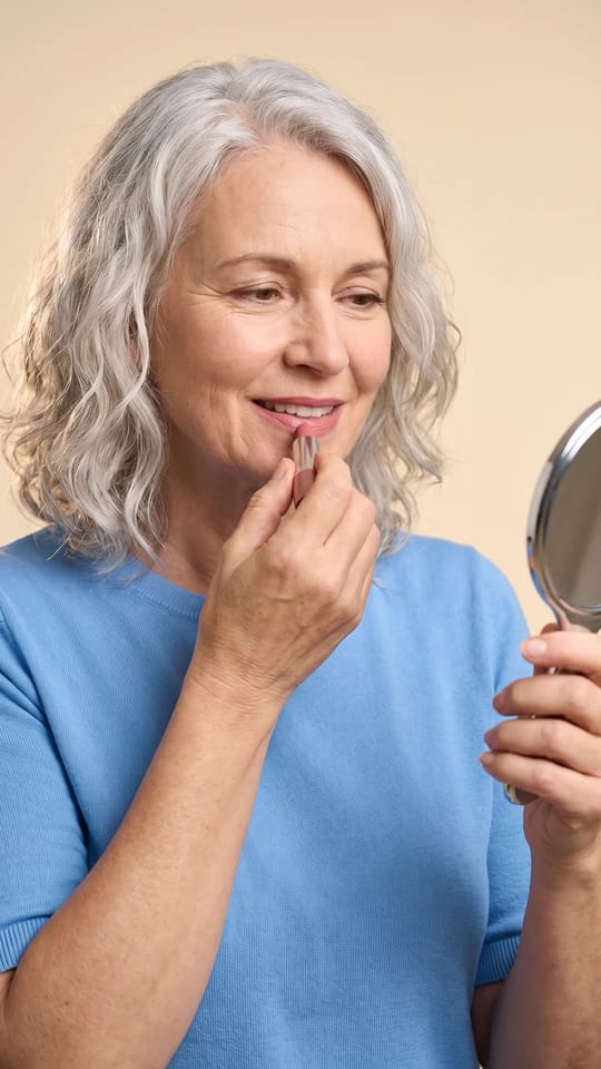 Mon rouge à lèvres à 55 ans : Pendant un cours, elle pose avec soin un rouge à lèvres rose doux devant un petit miroir à main, un maquillage naturel déjà en place, concentrée avec un petit sourire.