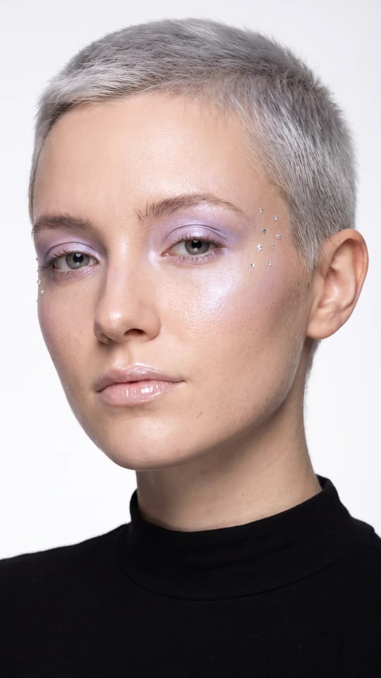 Éditorial céleste holographique : Éditorial céleste : fard opalescent lilas et nacre comme de la poussière de lune, highlighter holographique sur les pommettes et l'arcade, petits strass-étoiles aux tempes, peau translucide et lèvres perlées.