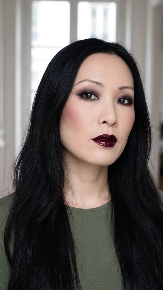 Gothique romantique glossy : Gothique romantique : lèvres cerise noire brillantes, yeux fumés anthracite et bordeaux, sourcils fins, peau porcelaine mate, blush bordeaux léger et petite aile de liner.